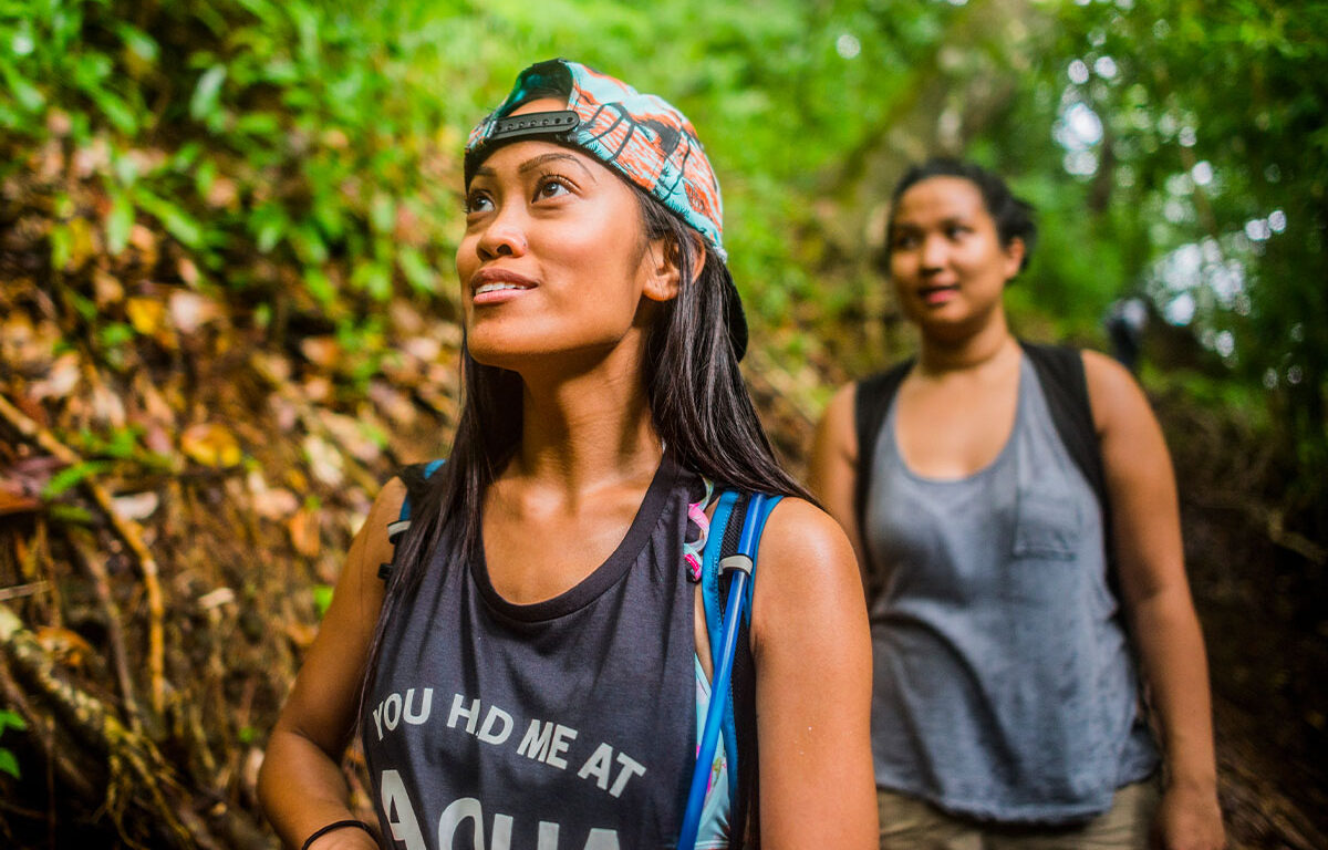 Two women hike along a Hawaii trial, looking up at the trees