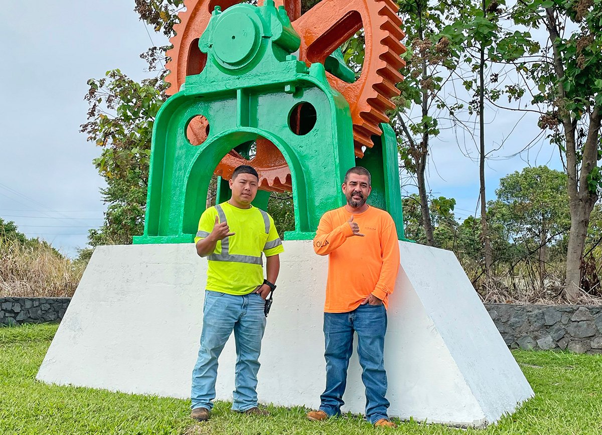 Two employees stand outside in front of a large green and orange statue of a gear. They are both smiling and holding hands up in a shaka towards the camera.